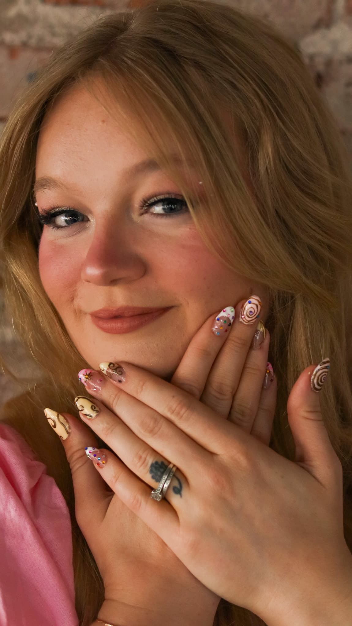 Kayla holding her hands near her face to show off her custom swirl and gem nail art, brick wall background